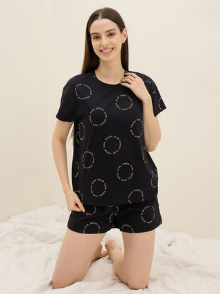 Women kneeling on a bed, wearing a black t-shirt and shorts set with a circular "couch club" print.