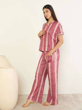 Women in a pink tie-dye pajama set, posing with one hand on her chest and the other in her pocket, with a white background and beige vases.