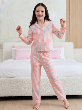 A cheerfully smiling girl stands in a pastel pink long-sleeved pajama set featuring vertical stripes in pink, light blue, and yellow, with an embroidered bear on the top.