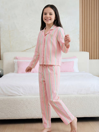A young girl stands smiling, wearing a pink, peach, and teal striped pajama set with a button-up top, long pants, and a small teddy bear emblem.
