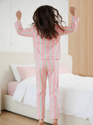 A woman in a striped pink, peach, and mint green long-sleeved pajama set jumps with her arms in the air, seen from behind.