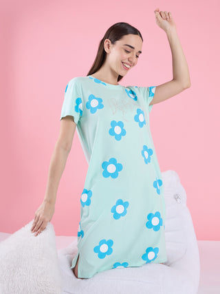A woman in a light blue nightgown with a floral print, sitting with her hand on a white pillow, with one arm raised.