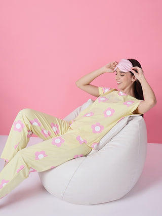 A woman in yellow floral print pajamas and a pink eye mask sits on a white beanbag chair, looking up and smiling.