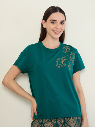 Women in a relaxed pose wearing a teal green t-shirt with gold leaf embroidery and matching patterned pajama bottoms.