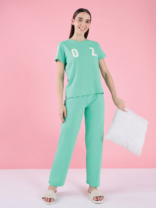 Woman in a mint green pajama set with white "02" print, holding a fluffy white pillow, posing with slippers on a pink background.