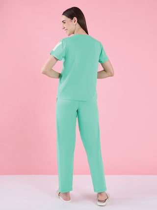 Woman in a mint green pajama set with white "02" print, posing with slippers on a pink background.