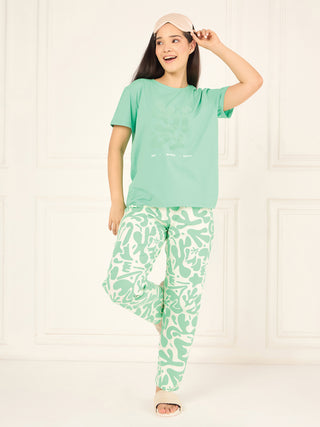 A woman poses playfully in mint green pajamas with a white abstract print and a sleep mask.
