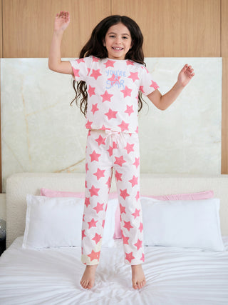A joyful girl in a white and pink star-print sleepwear set featuring "YOU'RE A STAR" messaging bounces happily on a bed.