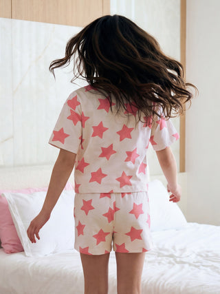 A young girl with dark, flowing hair is captured from behind, playfully turning while wearing a cream-colored short-sleeved pajama set featuring a cheerful pink star print.