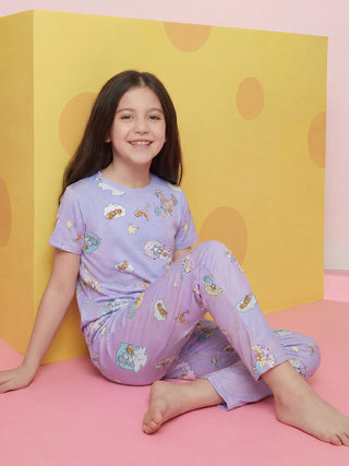 A smiling girl stands slightly turned, wearing a light purple short-sleeved and long-legged pajama set with a dreamy print of cartoon mice, cats, clouds, and stars.