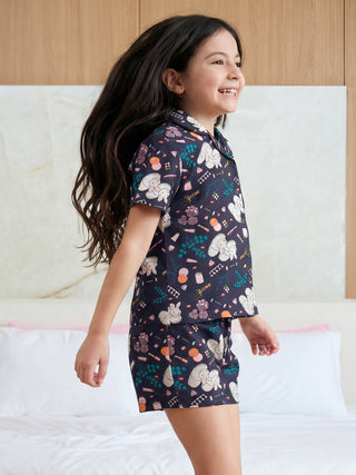 A happy young girl joyfully jumps in a dark blue short pajama set printed with playful bunnies, flowers, and cosmetic illustrations.