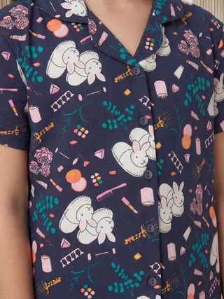 A young girl poses front-facing wearing a navy blue short-sleeved pajama top with a charming print of white bunnies, pink roses, candles, and cosmetic items among green foliage.