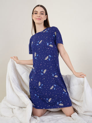 A woman kneels on a bed, wearing a short-sleeved, dark blue nightgown with a celestial floral print.