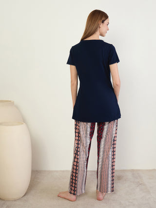 A woman stands wearing a navy blue t-shirt with decorative trim and patterned sleep pants featuring a geometric print in red, cream, and black.