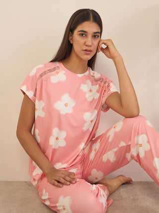 Woman seated in a relaxed cross-legged pose wearing a peach-pink short-sleeve sleepwear top with lace shoulder detailing and matching pyjama pants, featuring an all-over white floral print.