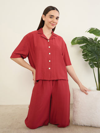 A woman in a red pajama set with pearl buttons, kneeling on the floor next to a fluffy cushion and a potted plant.