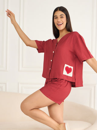 A woman in a red pajama set with a heart print stands with one leg bent and her arm extended, smiling.