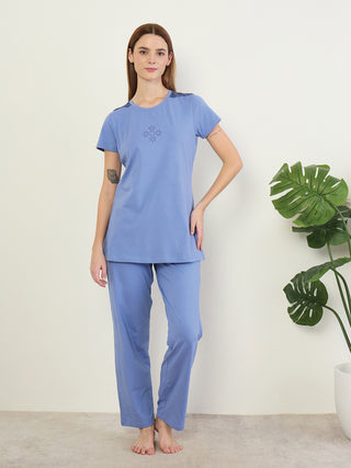 A woman stands wearing a blue pajama set with a floral print on the chest and a decorative detail on the shoulders.