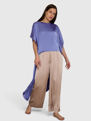 Women in a relaxed pose wear a flowing periwinkle tunic with side slits over beige satin wide-leg pants.