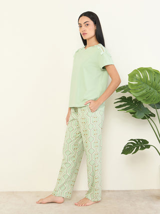 Women in a light green V-neck top with decorative shoulder accents and matching green patterned pants stand with one hand in her pocket.