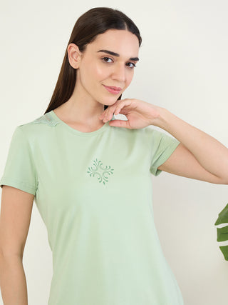 A woman poses wearing a pale green, short-sleeved t-shirt with a subtle lace detail on the shoulder and a decorative print on the chest.