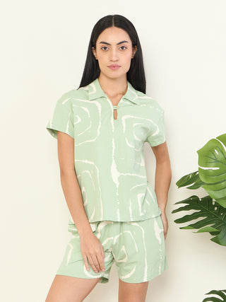 A woman in a light green, abstract-print sleepwear set, posing with hands on hips, stands beside a large monstera plant.