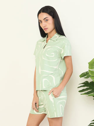 Women in a light green, swirl-printed sleepwear set are posing sideways with their hand in their pocket.