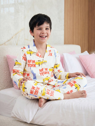 A cheerful boy happily sits cross-legged on a bed wearing cream-colored pajamas with a vibrant yellow taxi and 'NEW YORK' print.