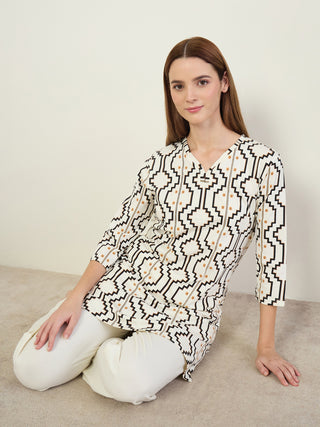 Women sitting down wearing a white tunic with a black and gold geometric print and white pants.