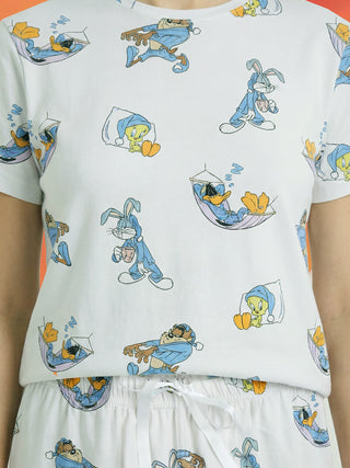 A woman stands wearing a white short-sleeved pajama set featuring a playful print of Looney Tunes characters like Bugs Bunny, Daffy Duck, Tweety, and Tasmanian Devil in various sleepy poses and blue sleepwear.