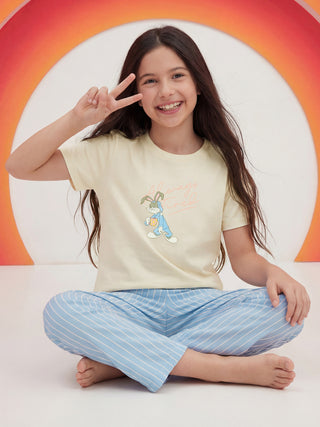 A young girl sits cross-legged, smiling and making a peace sign, in a pale yellow t-shirt with an 'Always Tired' rabbit print and light blue striped pajama pants.