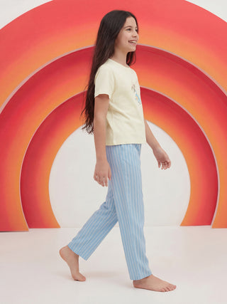 A smiling young girl walks barefoot in profile, wearing a light yellow short-sleeve pajama top with a subtle character print and light blue vertical striped pajama pants.