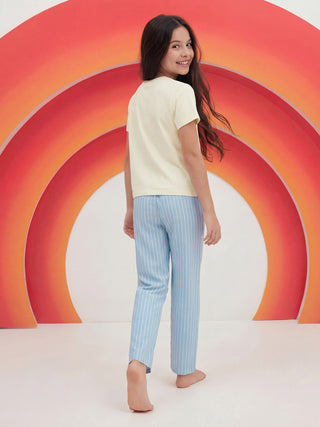 A young girl wearing a pale yellow short-sleeved sleepwear top and light blue vertically striped sleep pants smiles over her shoulder.