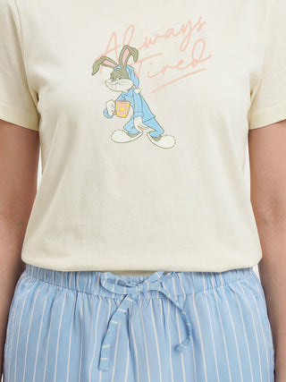 A female model stands wearing a cream short-sleeved sleepwear top featuring a sleepy Bugs Bunny graphic with pink "Always Tired" text, paired with light blue and white striped drawstring pajama bottoms.