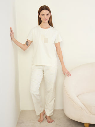 A woman stands in a cream-colored t-shirt and cream-colored patterned pajama pants.