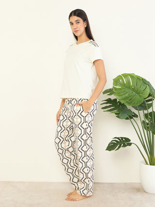 Women wearing a cream-colored t-shirt with embroidered shoulders and matching patterned pants, posing against a plain wall with a potted plant.