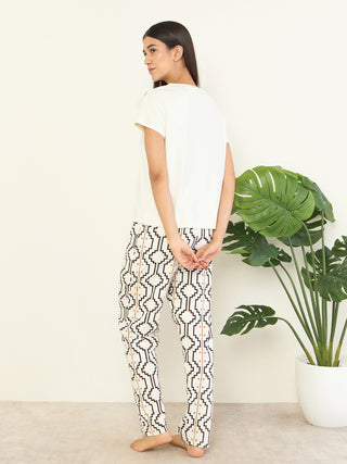 Women's cream t-shirt and patterned pants, standing with hands behind her back, a plant to her side.
