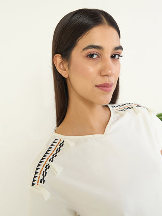 Women's cream t-shirt with embroidered shoulder detail and tassels, looking over her shoulder.