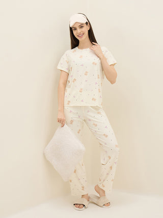 A young woman poses in ivory pajamas with a teddy bear print and a pink sleep mask, holding a fluffy white pillow.