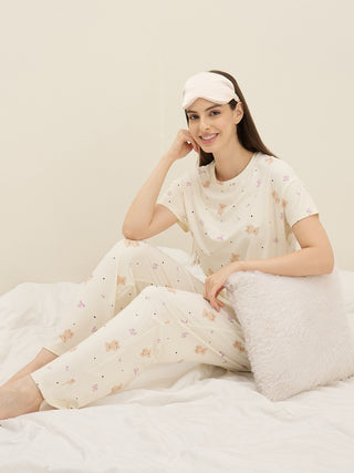 A woman sits on a bed wearing light cream pajamas with a teddy bear print and a pink eye mask.