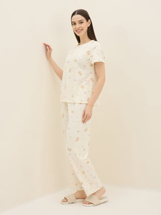 A woman in a cream-colored pajama set with a teddy bear print poses next to a wall.
