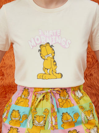 A model wears a cream short-sleeved t-shirt featuring a grumpy Garfield and "I HATE MORNINGS" text, paired with playful multi-colored pajama shorts patterned with various Garfield expressions.