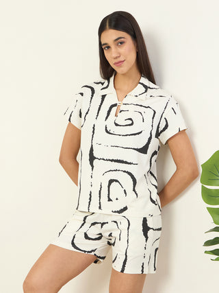 Women posing with hands behind their back, wearing a cream-colored pajama set with a black abstract swirl print.