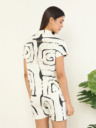 Women in a relaxed pose, wearing a cream-colored pajama set with a black abstract swirl print and a tropical graphic on the back of the shirt.