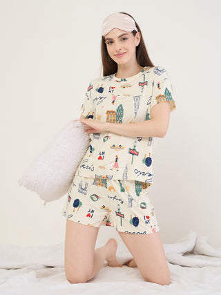 A woman is kneeling on a bed wearing light yellow sleepwear with a cityscape print and a pink eye mask.