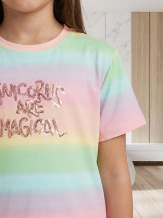 A young girl in a relaxed side profile wears a soft, short-sleeve pastel rainbow t-shirt with 'Unicorns Are Magical' spelled out in shimmering rose gold sequins.
