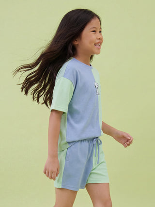 A smiling young girl with long, flowing dark hair walks while wearing a mint green and light blue color-block two-piece loungewear set featuring a small Snoopy print on the top.