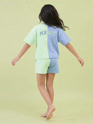 A young girl turns away from the camera, arms slightly outstretched, wearing a comfortable two-piece sleepwear set featuring a split design in pale green and periwinkle blue with "PEANUTS" printed across the back.