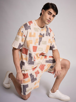A man in a beige t-shirt and shorts set with a geometric print, kneeling on a grey backdrop, wears white sneakers.