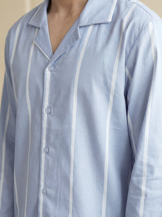 A man stands wearing a light blue pajama shirt with white vertical stripes and white piping along the revere collar and button-front.